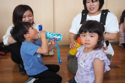 2組の親子が写っており、男の子が青い水稲を持って夢中で飲んでおり、女の子のお母さんふは黄色い水稲を手に持ち、女の子はカメラ目線でこちらを見ている写真