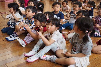 子ども達が床に体育座りをしており、両手をグーと握って小指だけ立てて、手遊びをしている写真