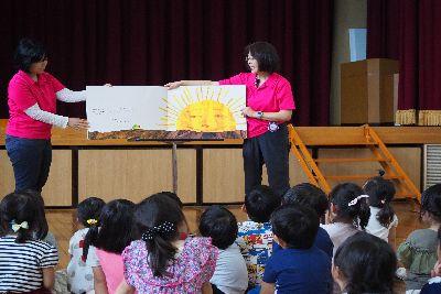 台の上に大きな絵本が広げられ、スタッフの女性二人で絵本の読み聞かせが行われており、沢山の子ども達が床に座って絵本に見入っている後ろ姿が写っている写真