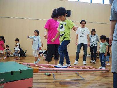 木のはしごの上を渡る男の子と、周りで見ているスタッフやお父さん、お母さんの写真