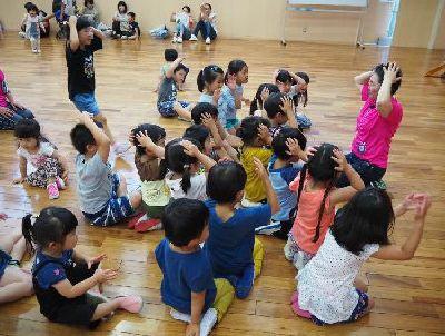 子供たちの前にいる女性スタッフと、子供たちが両手を頭においている写真