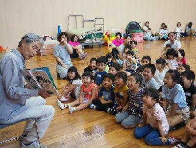 椅子に座った男性が、絵本の読み聞かせをしており、子供たちが笑顔で聞いている写真