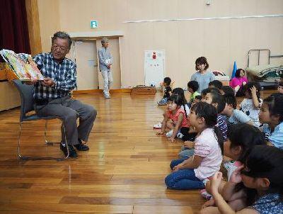 チェックシャツの男性が、子供たちの前で読み聞かせをしている写真