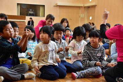 子どもたちが床に座っていて、左端にいる男の子がスタッフの女性の真似をして薬指を立てている写真