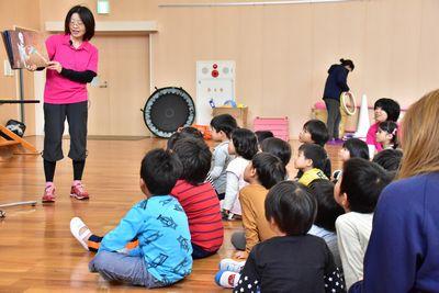 スタッフの女性が絵本の読み聞かせをしていて、子供たちが床に座って絵本を見ている写真