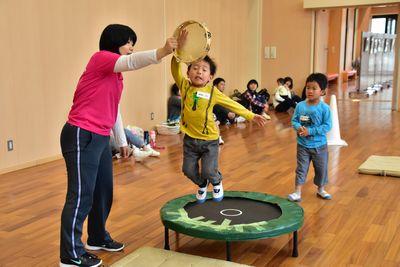 円形の小さなトランポリンを男の子が飛んで、横にいるスタッフが持っているタンバリンにタッチをしている写真