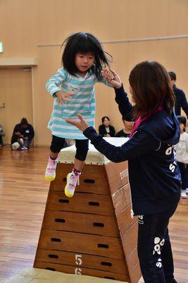 小さな女の子が6段の跳び箱から降りるのを女性スタッフが手を握って手伝っている写真