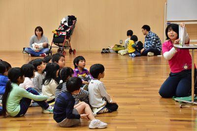 ピンクのシャツを着たスタッフが台の上に本を置きその横で本を読み、それを床に座って聞いている子供たちの写真
