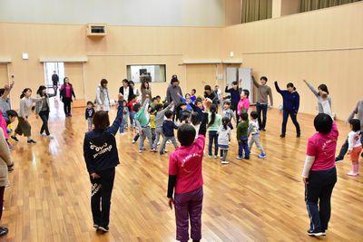 大人たちが右手を挙げ並んで大きな円を作りその中で子供たちが中央に集まっている写真
