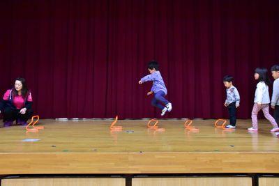 間隔をあけて置かれたオレンジ色のミニハードルを跳んでいる男の子と、順番を待っている子供たちの写真