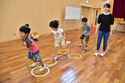 床に緑や白や黄色や赤の輪っかを並べて遊んでいる子供たちと1人の女性の写真