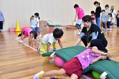 ボールの上に敷かれたマットに横たわっている子供、その後ろで四つん這いになっている子供、横で見守っている女性の写真