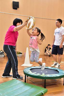 トランポリンの横でタンバリンを持っている女性と、トランポリンでジャンプしてタンバリンをたたいている女の子の写真