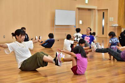 互いに向き合って床に座り、両足をそろえた状態でお互いの足の裏同士をくっつけて床から浮かせて両手を広げている親子の写真