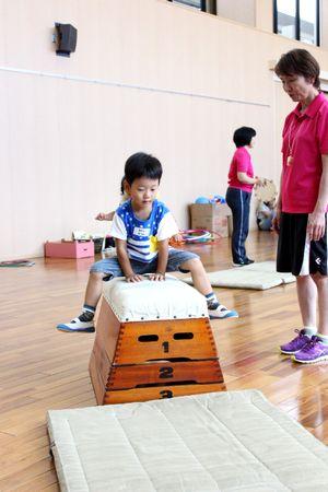 跳び箱にまたがっている男の子と横で見ている女性スタッフの写真