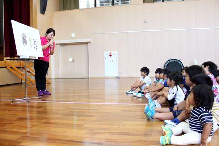 パネルの横で話をしている女性スタッフと床に座ってパネルシアターを見ている子供たちの写真