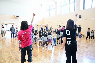子どもたちが円になって集まり片手を上に上げていて、周りの大人たちも一緒に片手をあげてガッツポーズをしている写真