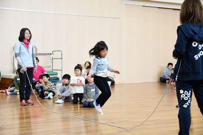 スタッフの女性が回す大繩を飛ぶ女の子と、座って見ている子供たちの写真