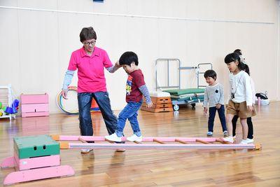 跳び箱に掛けられた木のはしごを、女性と手をつなぎながら渡る男の子と、その後ろに並ぶ子供たち3人の写真