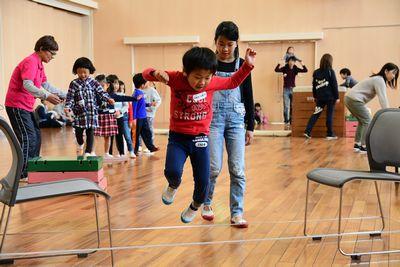 椅子と椅子の間に結ばれた紐を飛び越える男の子と、その後ろに立つ女の子の写真