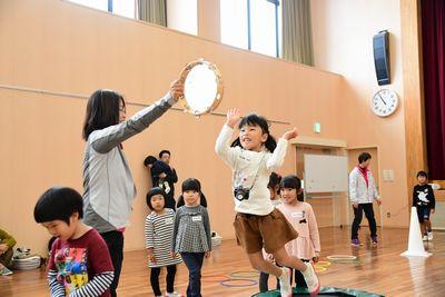 女性が持つタンバリンを、トランポリンでジャンプして触ろうとしている女の子の写真