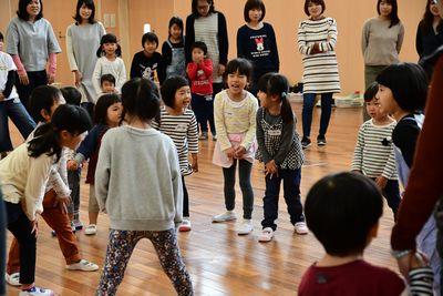 子供たちが輪になり、両手を片足の太ももの上に置きチクサクコールをし、その後ろで見ている他の子供たちや、お母さん達の写真
