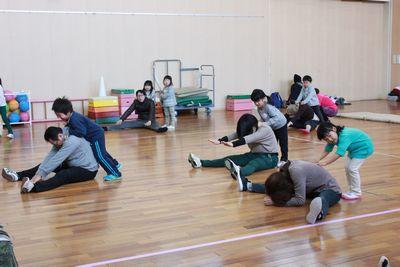 足を広げて床に座っている保護者の背中を子供たちが押している写真