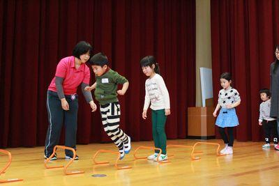 スタッフが見守る中、オレンジ色のミニハードルをジャンプしている男の子の写真