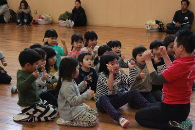 スタッフの周りに集まって座り、スタッフと同じ手の真似をしている子供たちの写真
