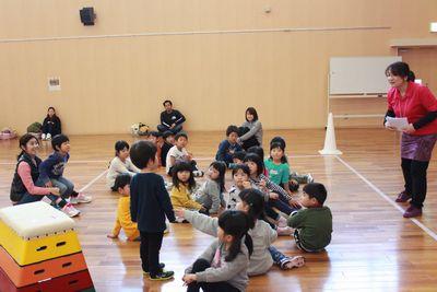 床の上に集合して座っている子供たちがおり、1人の男の子が立ち、周りの子供たちが立っている男の子の方を見ている写真