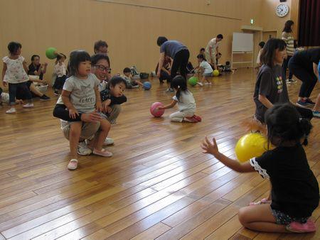 お父さんが小さい妹と弟をしゃがんで抱っこしており、お父さんの前に間をあけて座っているお姉ちゃんに向かって黄色いボールを投げている写真