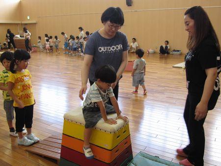 4段の跳び箱に男の子がまたがっており、お母さんが男児の側で危なくないように両手を側に置き、前ではスタッフの女性が見守っている写真