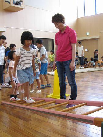 はしごが横に立てかけられており、その上を女の子が歩いており、ピンクのポロシャツを着た女性が見守っている写真