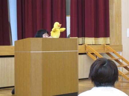 演台の後ろに女性が隠れて黄色いアヒルの指人形を手に付けて人形を使って話をしている写真