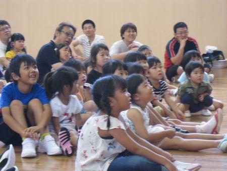 子ども達が床に座って絵本の世界に入って笑顔で見ており、後ろに座っている参加者のお父さんお母さんも笑顔で絵本を楽しんでいる写真