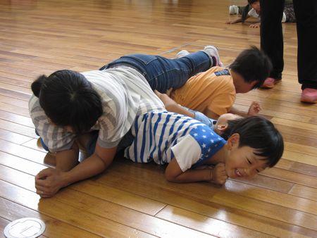 男の子二人が床の上に並んで寝転がっており、二人の男の子の上にお母さんが横になって男の子が重たそうに顔を歪めている写真