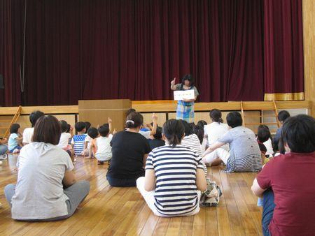 前方に絵本を広げて右手の人差し指を立てて絵本を読んでおり、その前に座っている子ども達もスタッフの女性と同じように人差し指を立て絵本を楽しんで見ている様子の写真