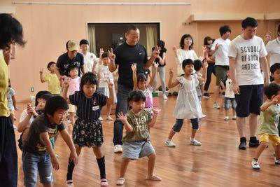参加者の親子が会場に広がっており、両足を広げて、肘をまげて両手をチョキにして挙げ、体操をしている写真