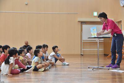 机の上に紙芝居を置いてスタッフの方がお話を読んでおり、子ども達が床に座って紙芝居を見入っている写真