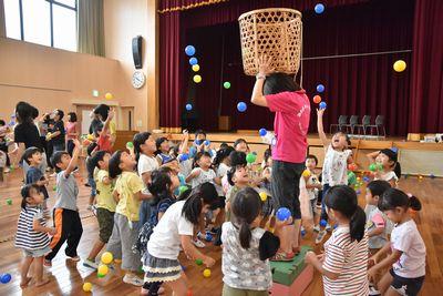 ピンクのポロシャツを着たスタッフが大きなかごを頭に乗せて跳び箱の上に立ち、子ども達がかごに向かってカラーボールを投げて玉入れをしている写真