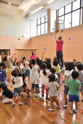 スタッフの女性が跳び箱の上に立って籠を両手を伸ばして高い位置に置いており、子ども達が籠に向かってカラーボールを投げている写真