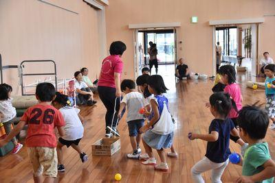 スタッフの方の女性が箱に紐を付けて、紐を引っ張って走っており、子ども達が拾ったボールを箱に入れようと追いかけている写真