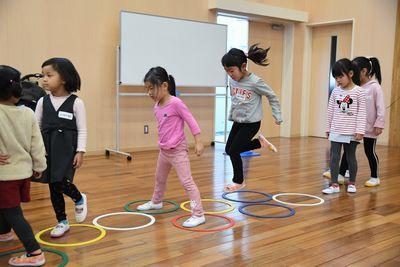 並べられた、カラフルな小さいフラフープのようなものに合わせて、けんけんぱをする女の子と、後ろに並ぶ子供たちの写真