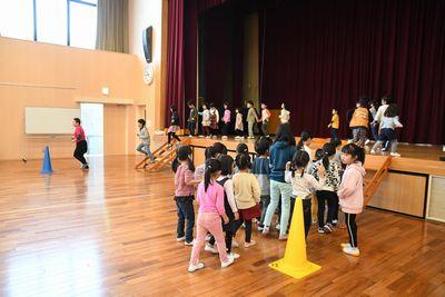 舞台の階段を上り、舞台を渡り、反対側の階段から降りる子供たちと、先頭で誘導する女性スタッフの写真