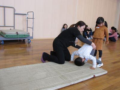 前回りをする男の子を女性が支え、その後ろに女の子が並んでいる写真