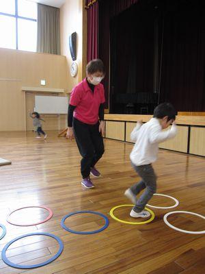 床に並べられた、カラフルな小さい輪に合わせて、けんけんぱをする男の子と、横に片足で立っている女性スタッフの写真