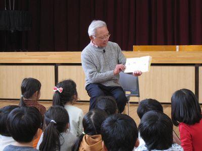 白髪の高齢の男性が、座っている子供たちの前で、読み聞かせをしている写真