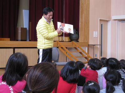 黄色いジャンパーを着ている男性が、座っている子供たちの前で読み聞かせをしている写真