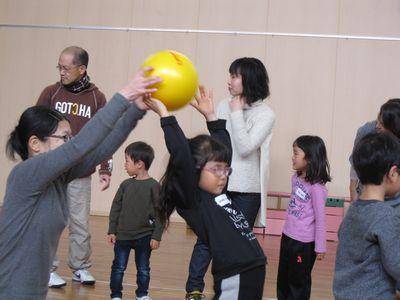 後ろの女性から、両手を高く上げて、後ろにそるような体勢でボールを受け取る女の子と、列を作る親子の写真