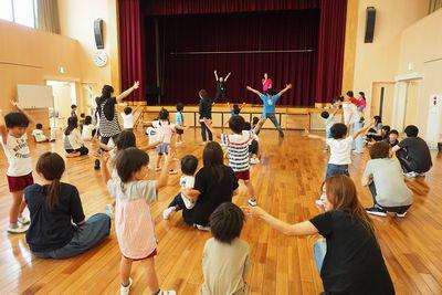 指導者が舞台上に立ち、その動きに合わせて子供たちが両手を上げて広げている写真